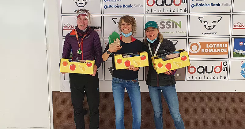 Drie vrouwen die glimlachen en gele dozen met fruitmotieven vasthouden, staande voor een sponsorachtergrond tijdens een evenement in een zaal. Ze lijken iets te vieren of prijzen in ontvangst te nemen.